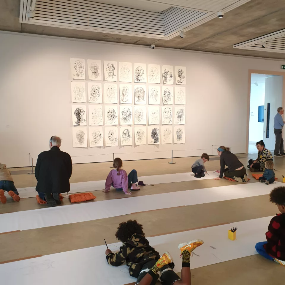 Children lying on the floor of the gallery drawing on large strips of paper.