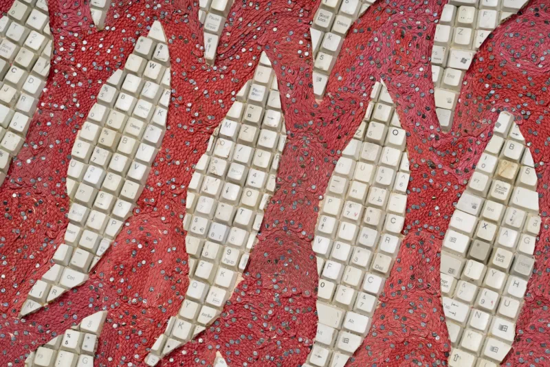 A colour photograph of Elias Sime's concave floor based sculptural piece, that is made from computer keys in leaf like shapes and red woven reclaimed electrical wires.