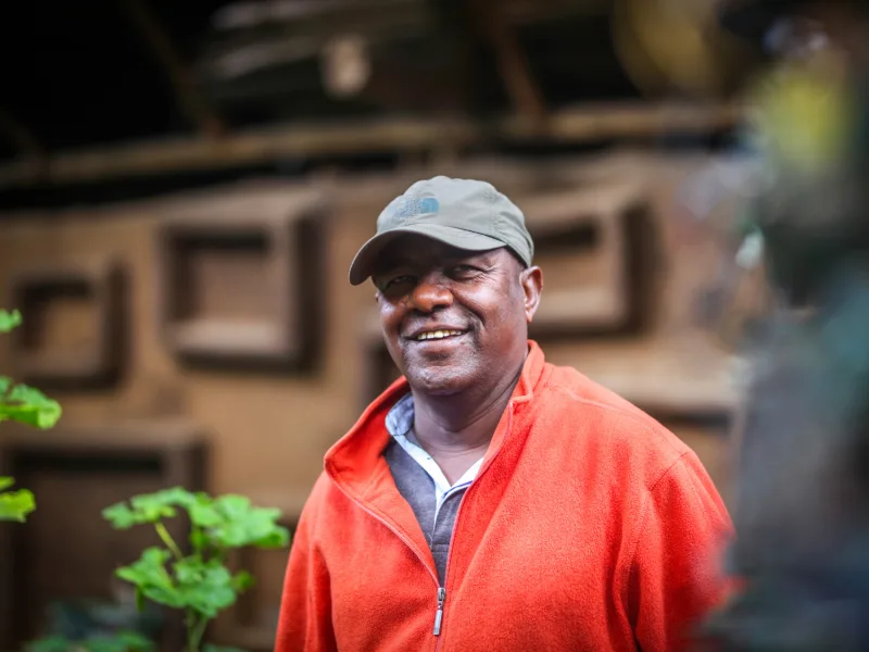 A colour photograph of artist Elias Sime, wearing a bright orange jacket and cap, smiling.